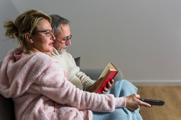 Aged woman with TV remote watching TV and man reading book on settee