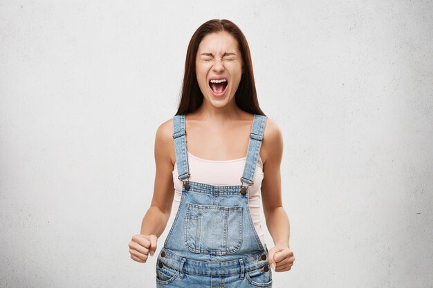 Aggression, negative human reaction and attitude. Indoor shot of angry stressed out young female screaming out loud with mouth wide opened, eyes closed and fists clenched, having mad and furious look