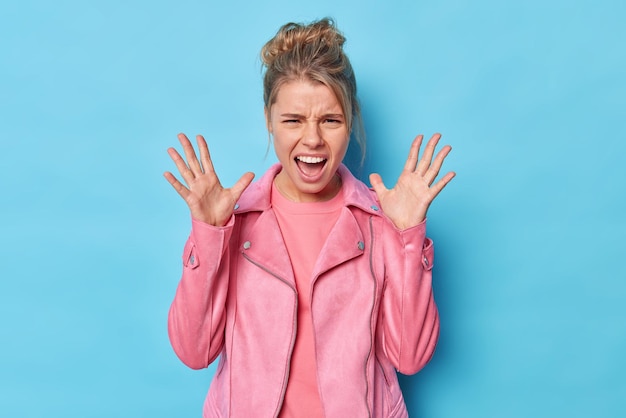 Free Photo aggressive displeased young woman raises palms screams angrily expresses negative emotions keeps mouth opened wears pink jacket poses against blue studio background. i cannot stand this anymore