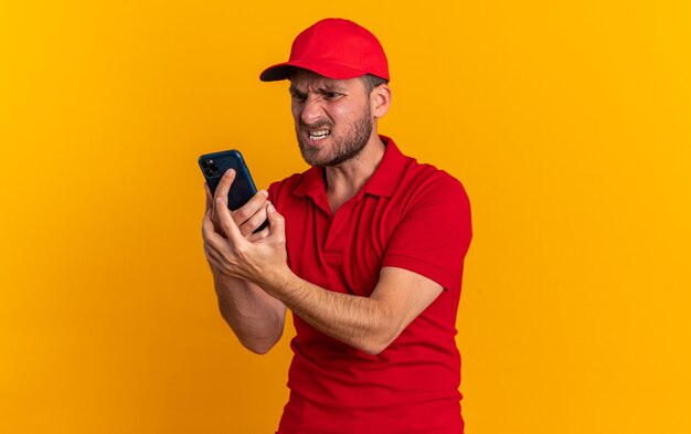 Aggressive young caucasian delivery man in red uniform and cap standing in profile view holding and looking at mobile phone isolated on orange wall with copy space