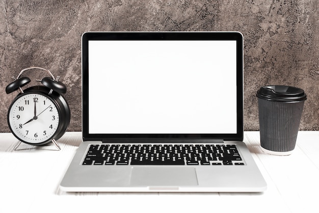 Free photo alarm clock and disposable coffee cup with an open laptop on white desk