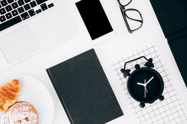 Alarm clock; laptop; cellphone; eyeglasses; baked croissant and buns on plate and diary over white background