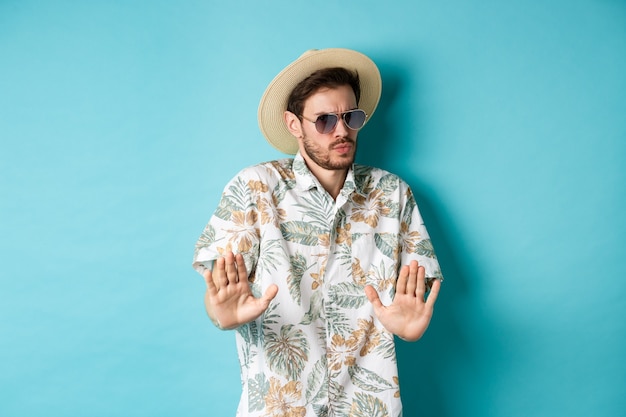 Alarmed tourist asking to stay away, step back from something cringe, showing rejection gesture, standing in straw hat and hawaiian shirt, blue background.