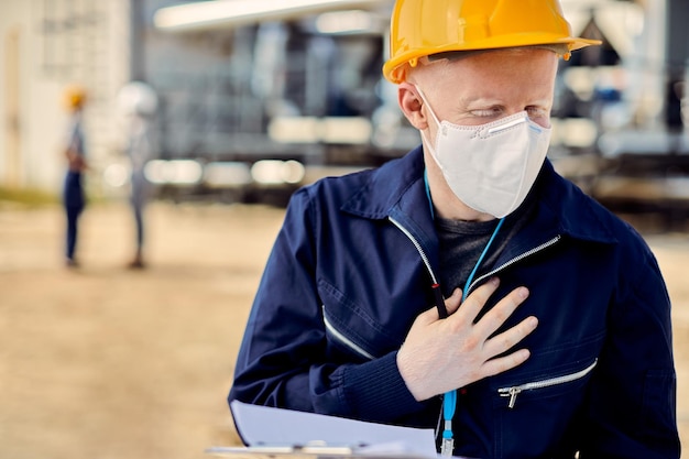 Albino construction worker with face mask holding his chest in pain while working outdoors