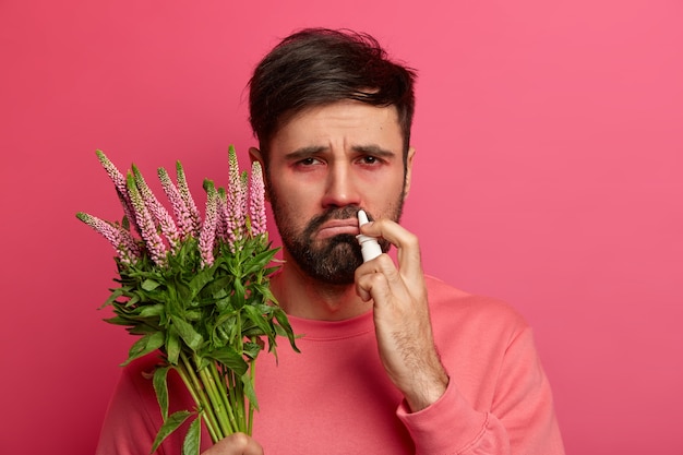 Free Photo allergic bearded man holds plant, uses nasal drops to cure sneezing, has displeased face expression, reaction on allergen, cureses seasoanl disease, follows advice of allergist. health care concept