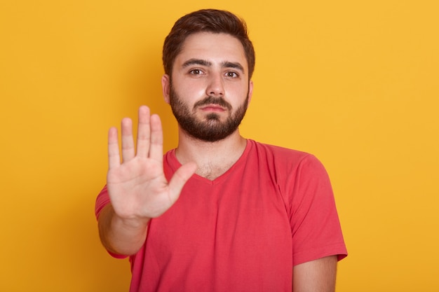 Free photo Сalm bearded young man wearing red casual t shirt standing with stop warning gesture hand and looking at camera with serious face