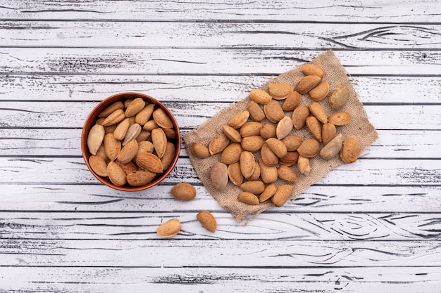 Almonds in the ceramic bowl near the other almond on sackcloth on white wooden surface top view