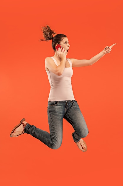 Always on mobile. Full length of pretty young woman taking phone while jumping against red studio background.