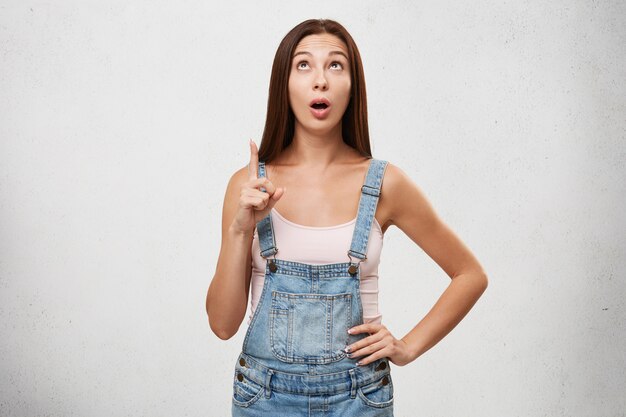 Amazed astonished cute teenage woman wearing stylish jumpsuit looking up in fascinating and pointing index finger, indicating something curious and shocking.