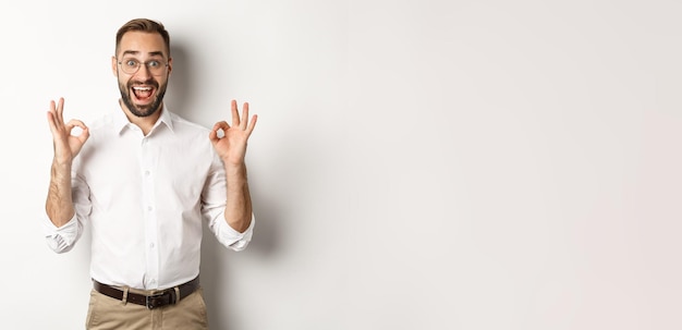 Free photo amazed entrepreneur showing okay sign and looking happy satisfied with product standing over white b