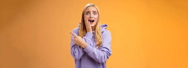 Free photo amazed excited overwhelmed young blond girl in hoodie press palm cheek screaming astonished pointing right seeing famous super star cannot hold joyful positive emotions orange background