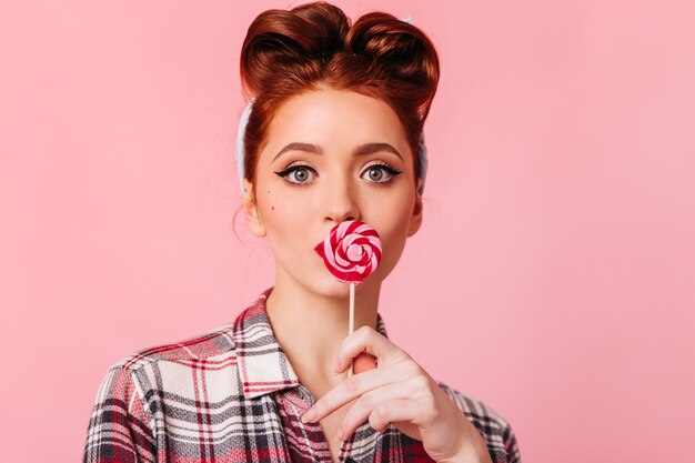 Amazed ginger girl in checkered shirt looking at camera. Studio shot of pinup woman with candy isolated on pink space.