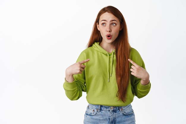 Amazed ginger girl gasp and say wow, pointing at herself and looking at logo aside, self-promoting, asking to pick her, wants to volunteer, white background.