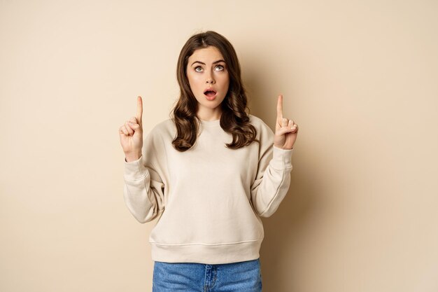 Amazed girl checking out banner announcement, reading promo text upwards, pointing fingers up, standing over beige background