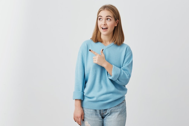 Amazed girl with opened mouth looking with dark eyes aside pointing with index finger and advertising something. Young woman posing  with copy space for text or promotion