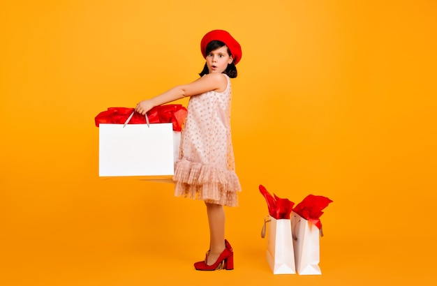 Amazed kid in red beret and moms shoes standing on yellow background Studio shot of cute child with shopping bags