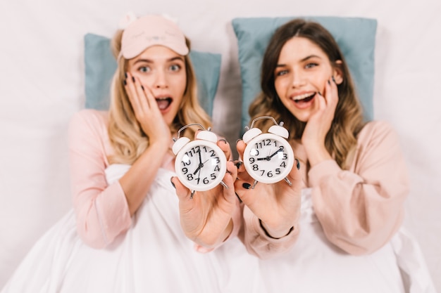 Amazed ladies posing in bed with clocks