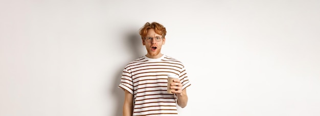 Free photo amazed redhead man in glasses holding coffee cup and staring at camera with complete disbelief stand