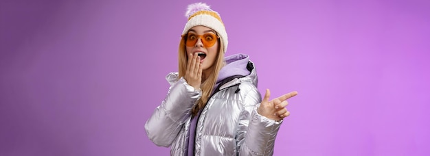 Free photo amazed speechless surprised attractive stylish blond girlfriend in sunglasses silver glittering jacket hat drop jaw impressed widen eyes shocked pointing left astonished purple background