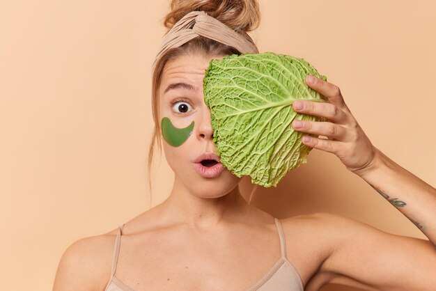 Amazed surprised woman holds breath feels amazed holds fresh cabbage over face applies hydrogel patches under eyes to remove wrinkles undergoes beauty procedures isolated over beige background