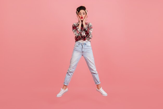 Amazed woman in jeans jumping on pink background. Pretty pinup girl in checkered shirt expressing surprised emotions.