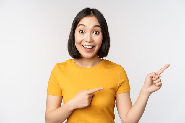 Amazed young asian woman showing advertisement aside pointing fingers right at promotion text brand logo standing happy against white background