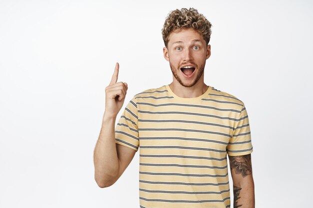 Amazed young blond man pointing finger up guy screams from excitement while showing awesome announcement recommending store with sale white background