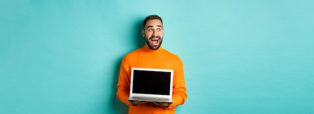 Free photo amazed young man in orange sweater looking at upper left corner showing laptop screen promo offer st