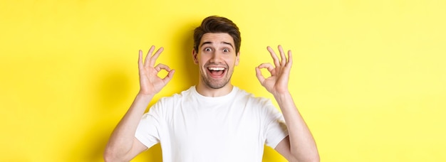 Free photo amazed young man showing ok signs and smiling recommending something good standing over yellow backg