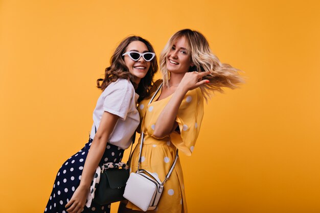 Amazing curly girl in white sunglasses having fun with best friend. Inspired sisters in trendy clothes posing on orange.