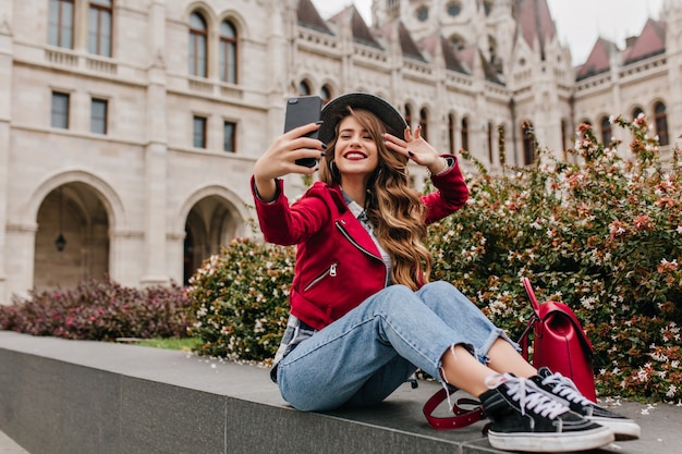 Amazing curly woman in black shoes making selfie in front of historical building