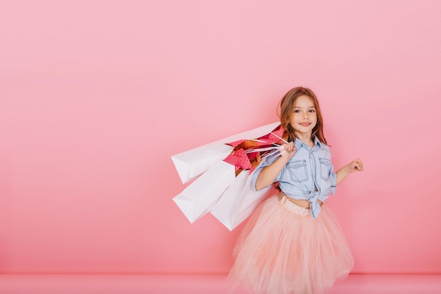 Free photo amazing cute little girl with long brunette hair walking with white packages isolated on pink background. little happiness, expressing true positive emotions to camera. place for text