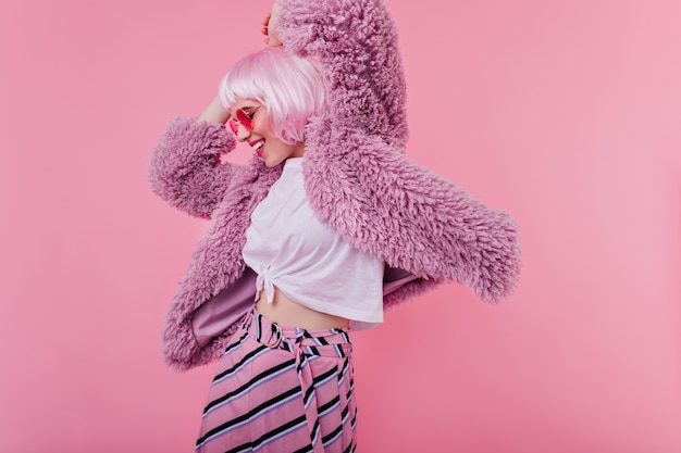 Free photo amazing girl in purple jacket having fun during indoor photoshoot.  cute female model in sunglasses and pink peruke dancing and laughing