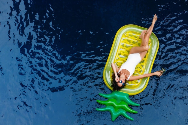 Free Photo amazing girl in sunglasses lying on pineapple mattress. outdoor photo of lovely tanned female model in swimsuit relaxing in pool.