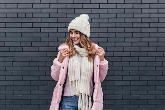 Amazing girl with beautiful smile enjoying photoshoot in cold morning. Fashionable blonde female model in coat and knitted hat posing near bricked wall.