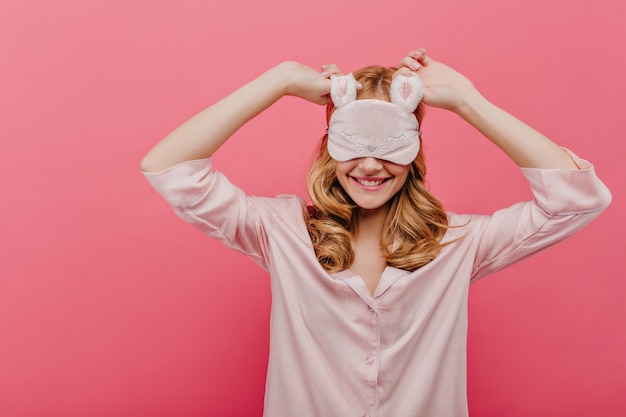 Free photo amazing girl with curly hair wears eyemask and laughing in weekend morning. blissful white female model in silk night-suit fooling around on pink wall.