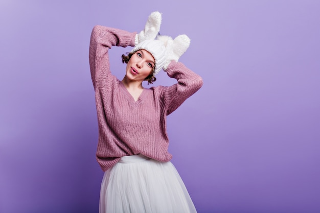 Free photo amazing girl in woolen gloves fooling around  indoor photo of charming female model in knitted hat and white skirt making funny faces on purple wall.