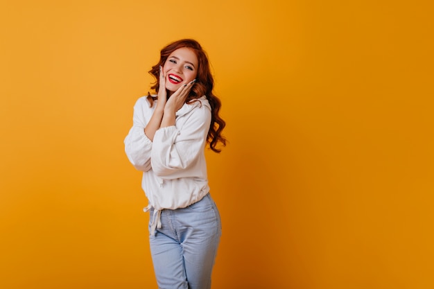 Amazing long-haired woman smiling on yellow wall. Gorgeous ginger female model posing in white sweater.