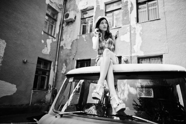 Free photo amazing long legs with hig heels girl wear on hat sitting on roof of old retro minivan and listening music from mobile phone