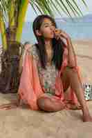 Free photo amazing sexy tan asian woman posing on paradise tropical beach under the pam tree, sitting on white sand , relaxing and enjoying holidays. boho dress with embroidery. bali.