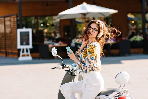 Amazing stylish girl with phone on scooter waiting for boyfriend who went for coffee in cafe