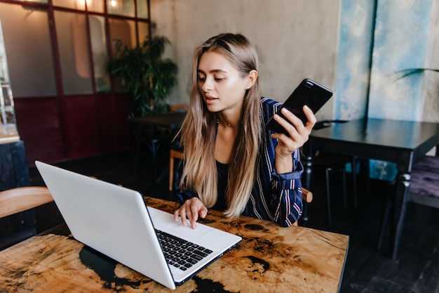 Amazing white girl with smartphone in hand working with laptop. Appealing female freelancer sitting in cafe.