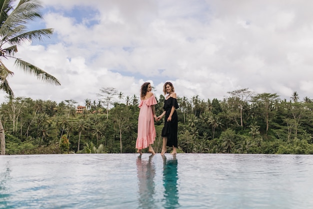 Free Photo amazing woman in long pink dress standing beside lake. charming ladies holding hands near outdoor pool with forest