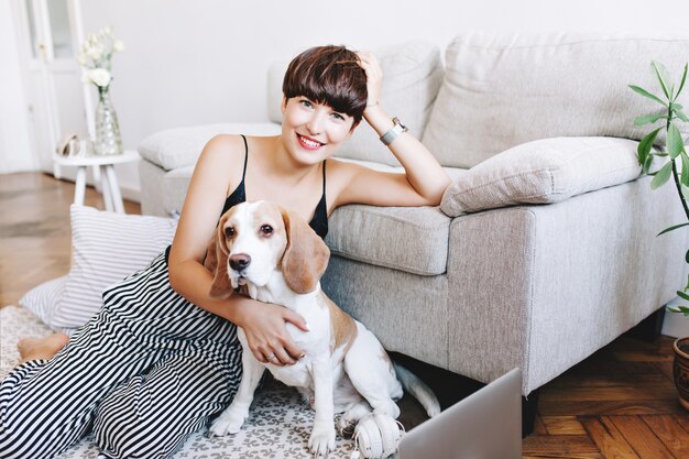 Amazing young woman wears striped pants and wristwatch posing on the floor while playing with beagle dog