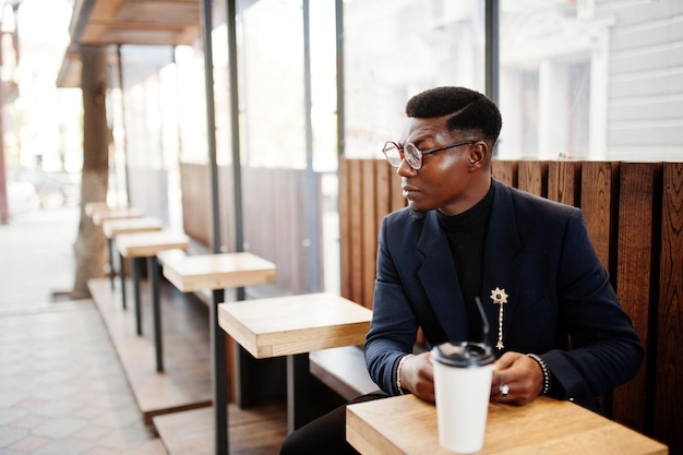 Amazingly looking african american man wear at blue blazer with brooch black turtleneck and glasses posed at street Fashionable black guy with cup of coffe looking at his phone