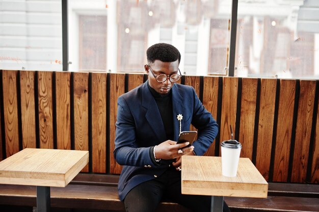 Amazingly looking african american man wear at blue blazer with brooch black turtleneck and glasses posed at street Fashionable black guy with cup of coffe looking at his phone