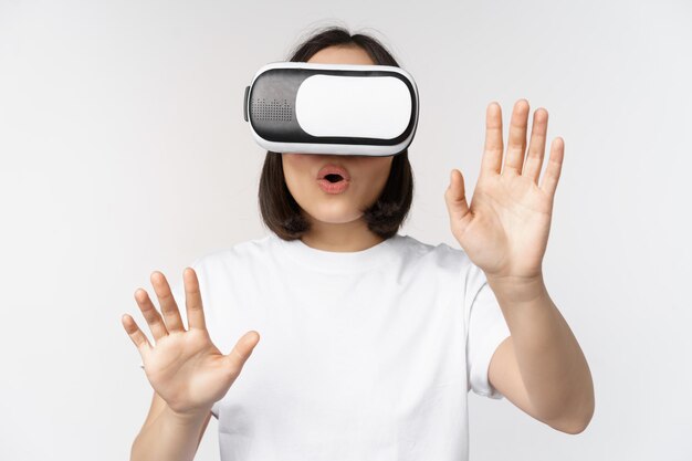 Amused asian girl using VR glasses virtual reality headset and reaching hands into empty space touching smth augmented standing over white background