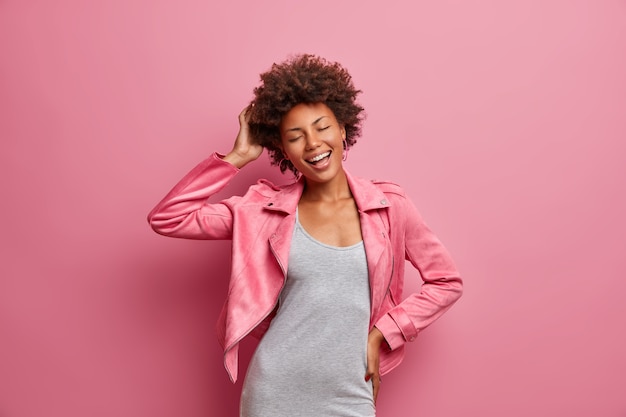 Amused carefree young Afro American woman with closed eyes, enjoys awesome day off, dressed in stylish clothes, smiles broadly