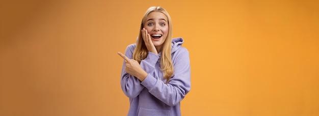 Free photo amused excited speechless happy young blond woman 25s in hoodie cannot believe luck happiness pointing right amazed smiling broadly touch cheek surprised receive awesome unexpected gift