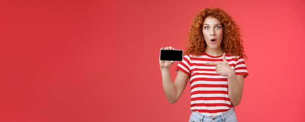 Free photo amused fascinated cute redhead curly girl adore playing games hold smartphone horizontal show thumb